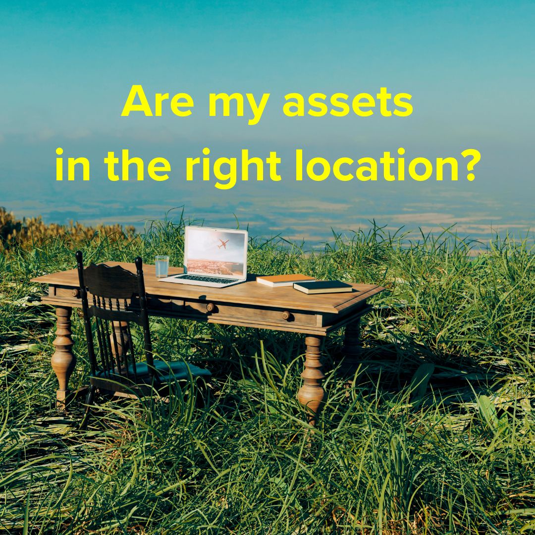 Title: 'Are my assets in the right location?' in yellow on top. Photo of a brown wooden desk and a black chair in tall grass at the top of a hill. On the desk are a laptop, two books, and a glass of water.
