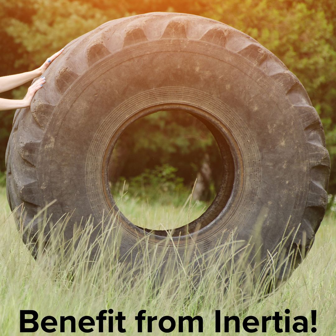 Woman pushing a huge tractor tire down a gentle slope in a field with tall grass in front of a wooded area. Words at the bottom on black text: 'Benefit from Inertia!'