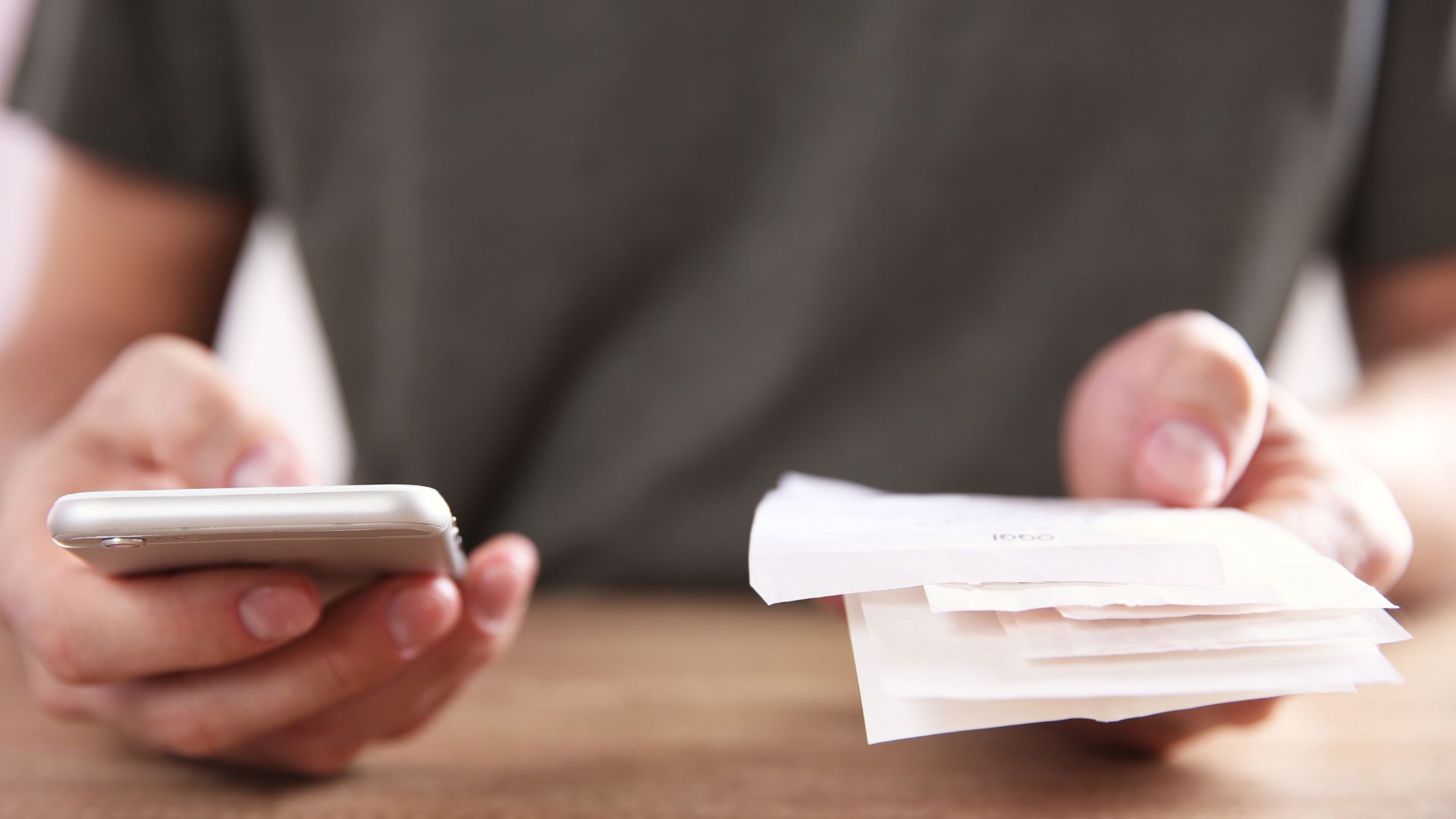 Photo of someone with their hands on a wooden table. They're holding their phone in their right hand and paper receipts in their left.