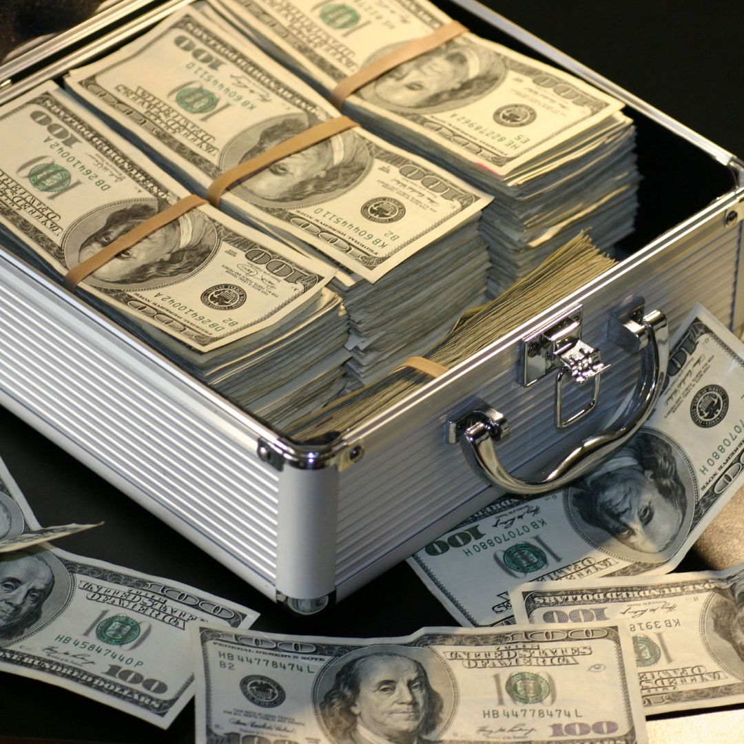 Silver metal case with stacks of hundred dollar bills in rubber bands. Some of the bills are lying around on a black table outside the case.