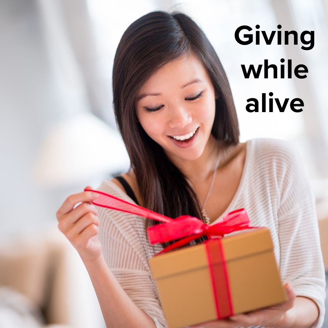 Title 'Giving while alive' in black font. A woman excitedly opens a gift with a red bow.