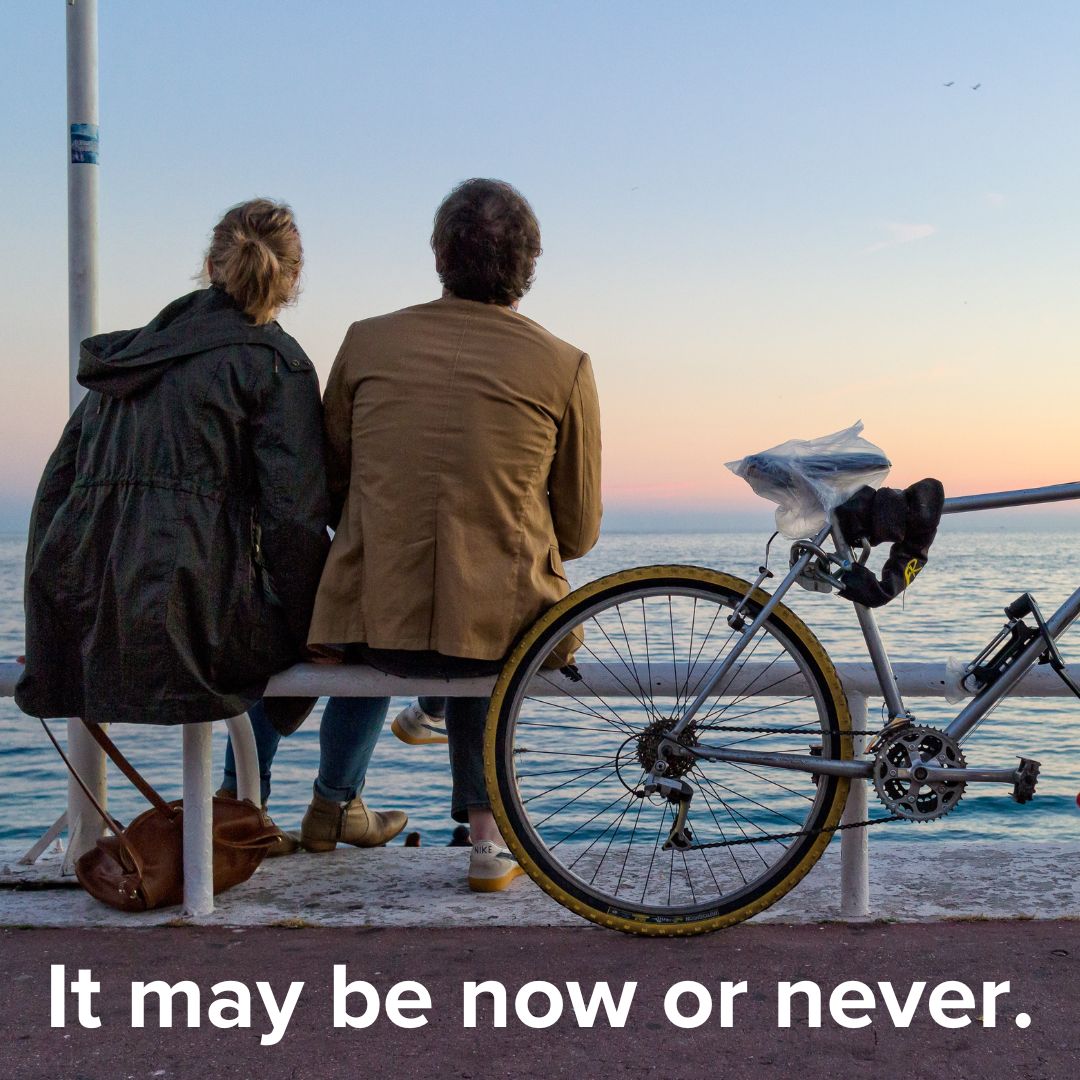 Title 'It may be now or never.' in white on bottom. The photo is of a couple sitting on a bench overlooking the water at dusk. A bicycle leans up against the bench.