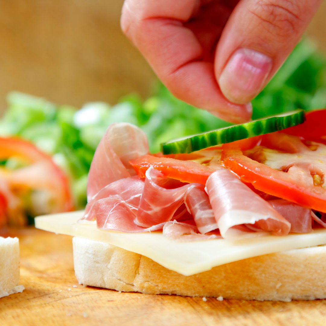 A hand adding a cucumber slice to the makings of a sandwich, complete with bread, cheese, meat, and tomato. Lettuce appears to be in the background. Another piece of white bread sits nearby.