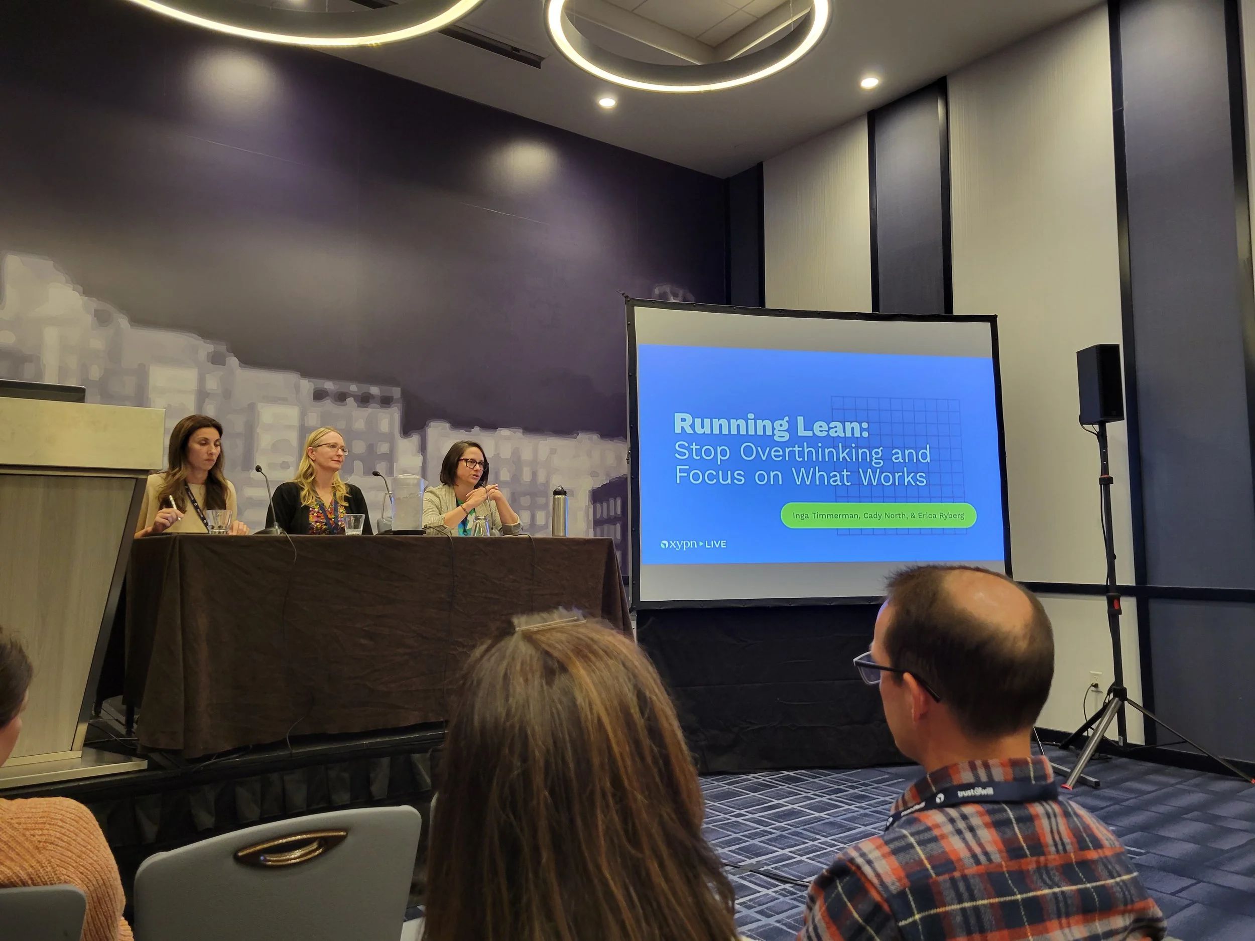 Photo of Inga Timmerman, Erica Ryberg, and Cady North presenting to a crowd in a large hotel room. The presentation title on a screen is 'Running Lean: Stop Overthinking and Focus on What Works.'