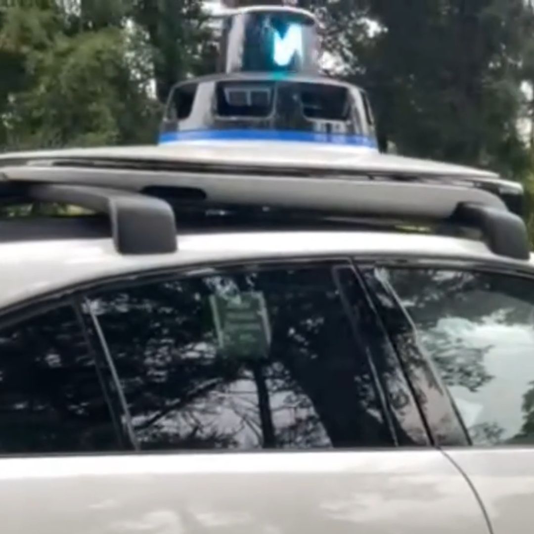 Photo of a white self-driving vehicle with an attachment on top. The attachment appears to be a large, blue light on top of a white disk.