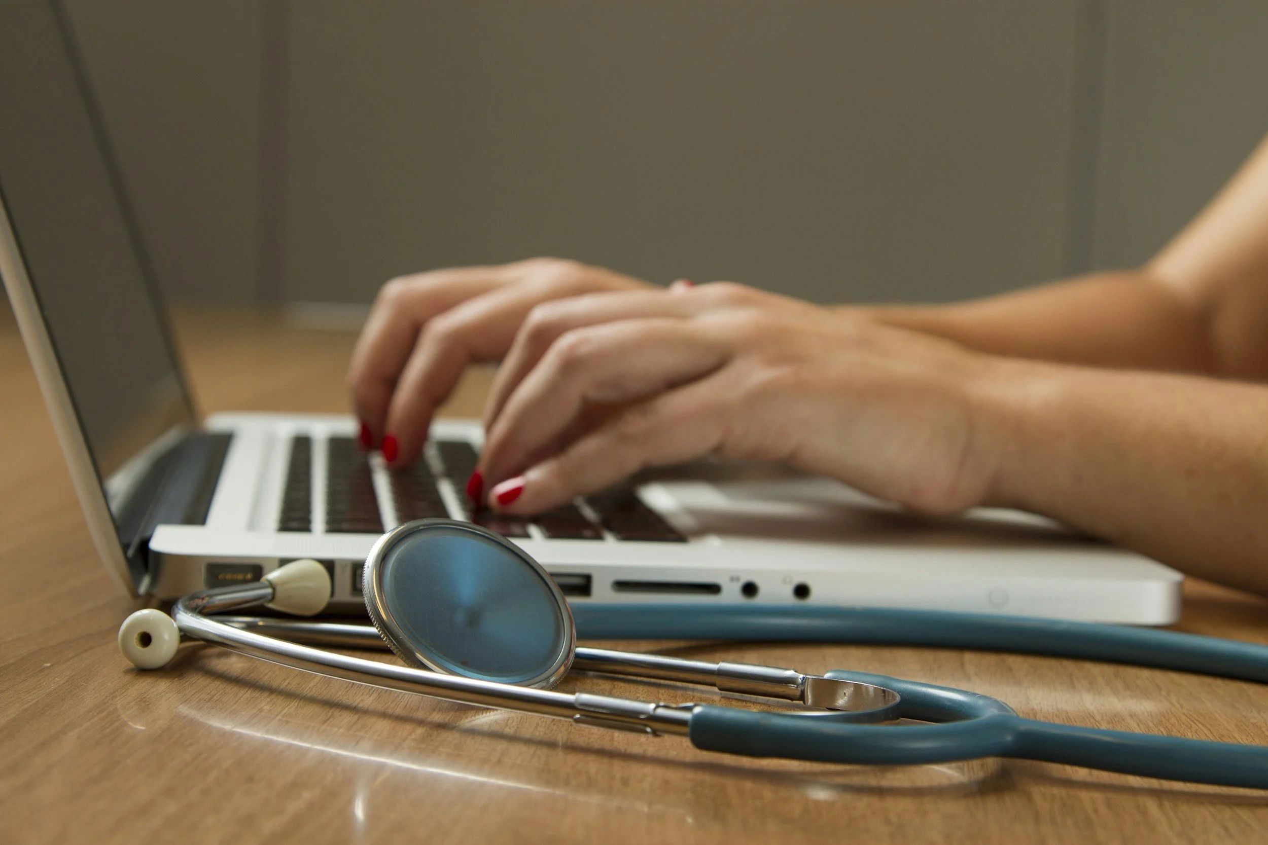 Someone with red nail polish typing on a computer. A stethoscope lies on a wooden table next to the computer.