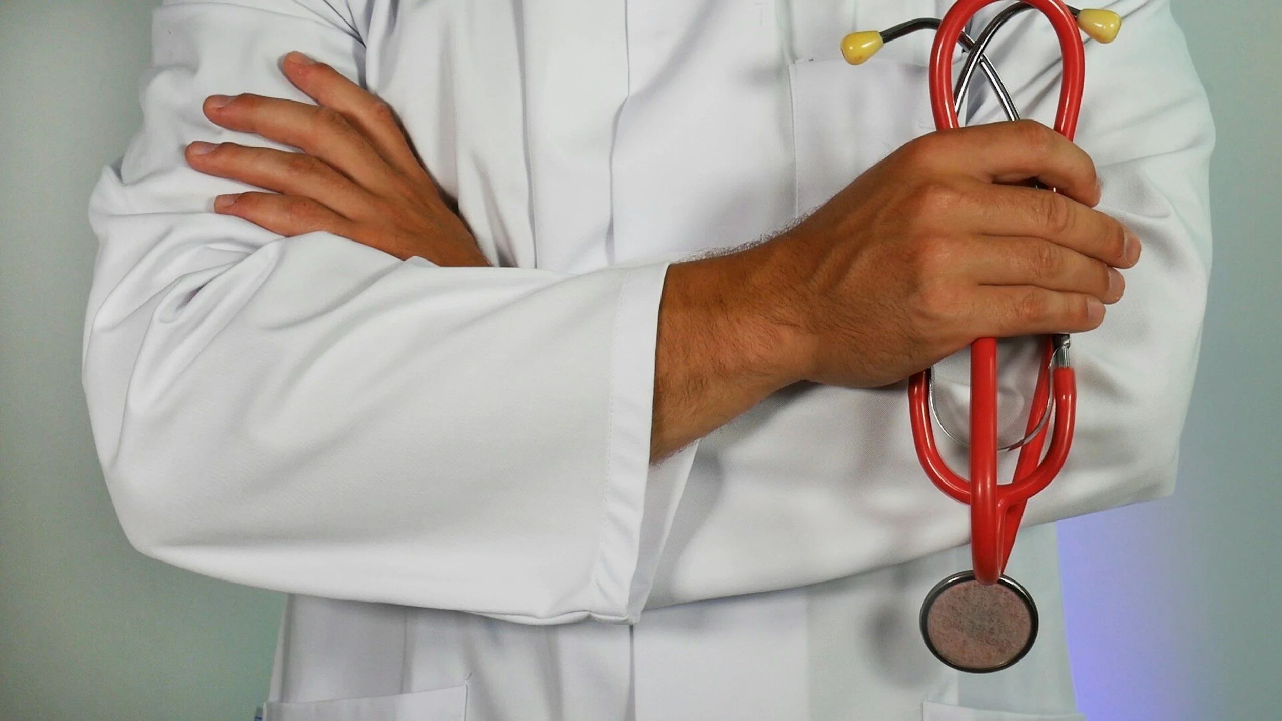 Photo of a doctor in a white coat is standing with arms crossed. One hand holds a stethoscope.