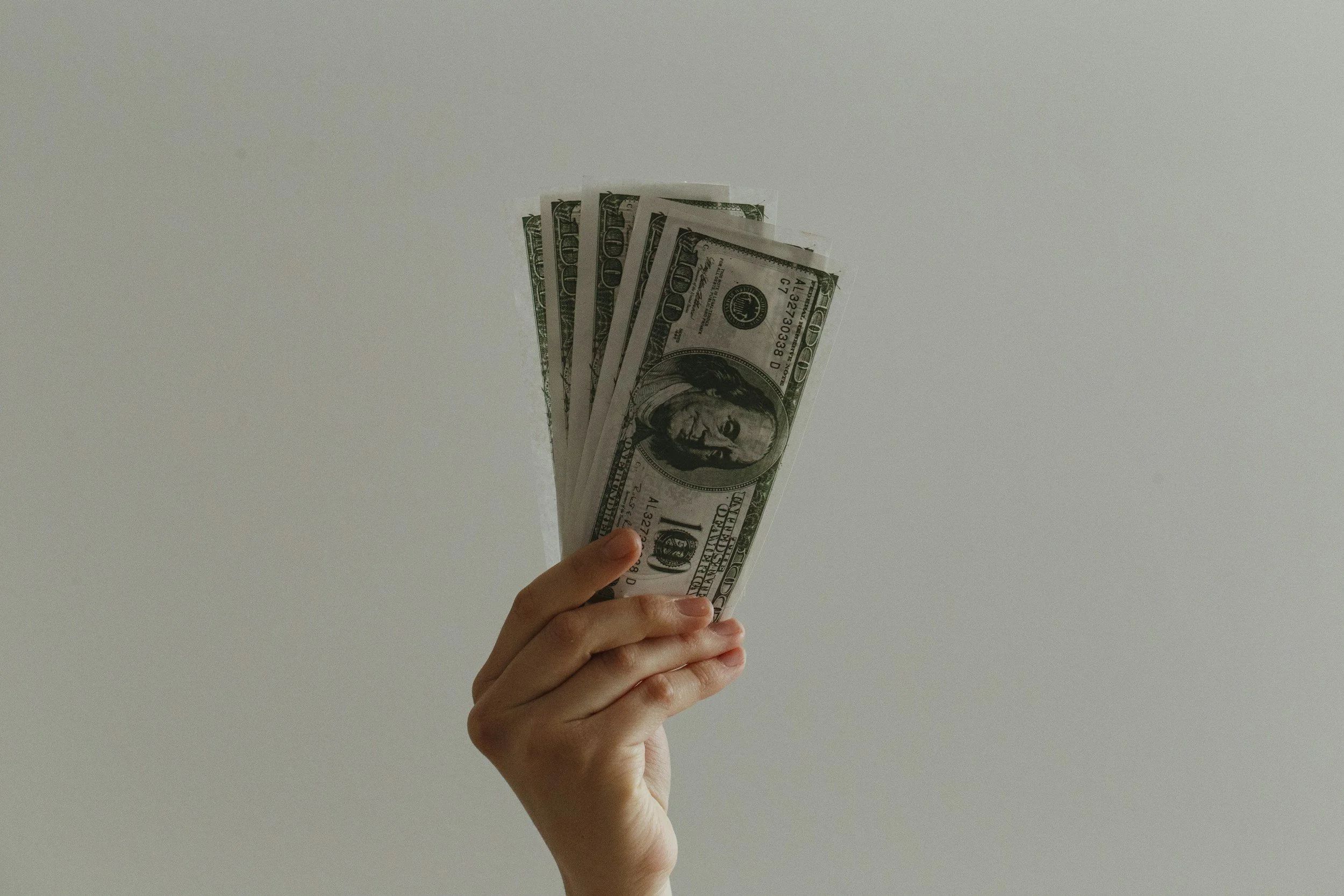 A hand holding up five $100 bills. The background is gray.