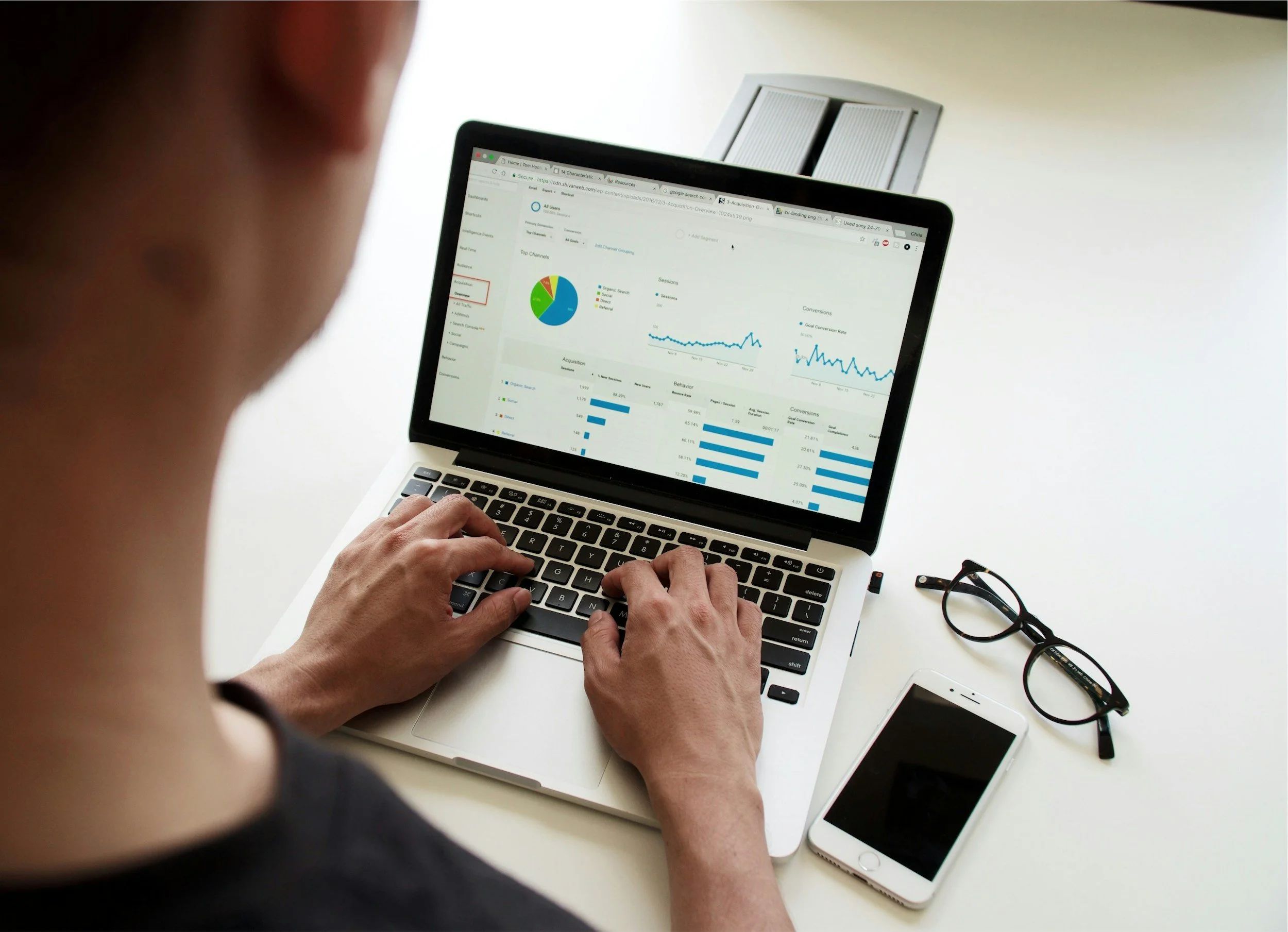 A photo of someone sitting at the desk with their back to the camera. They're logged into what appears to be a personal financial dashboard. There are a phone and glasses on what appears to be a conference room table.