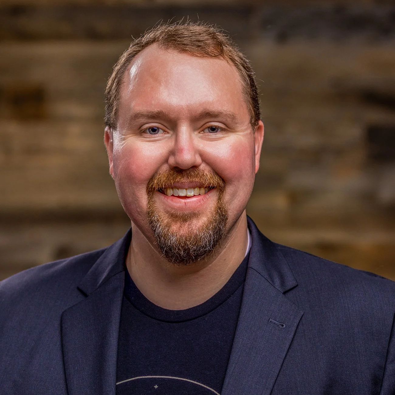 Kevin Estes portrait. Smiling and wearing a dark gray suit and black shirt in front of a dark brown background.