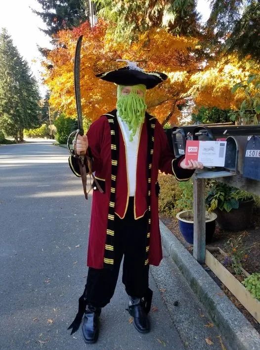 Kevin Estes dressed like a pirate in a red coat, fake sword, crocheted tentacle green mask, tri-point hat, and holding a voter's ballot in front of a mailbox and orange-colored trees.