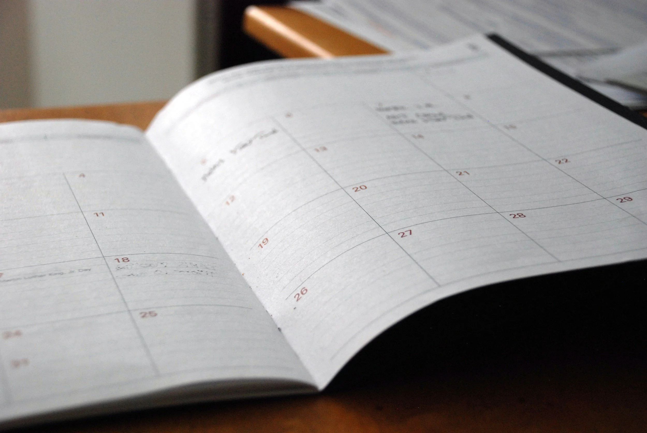 A photo of a paper calendar for a single month with entries on a few of the days. The calendar rests on a wooden desk with other items.