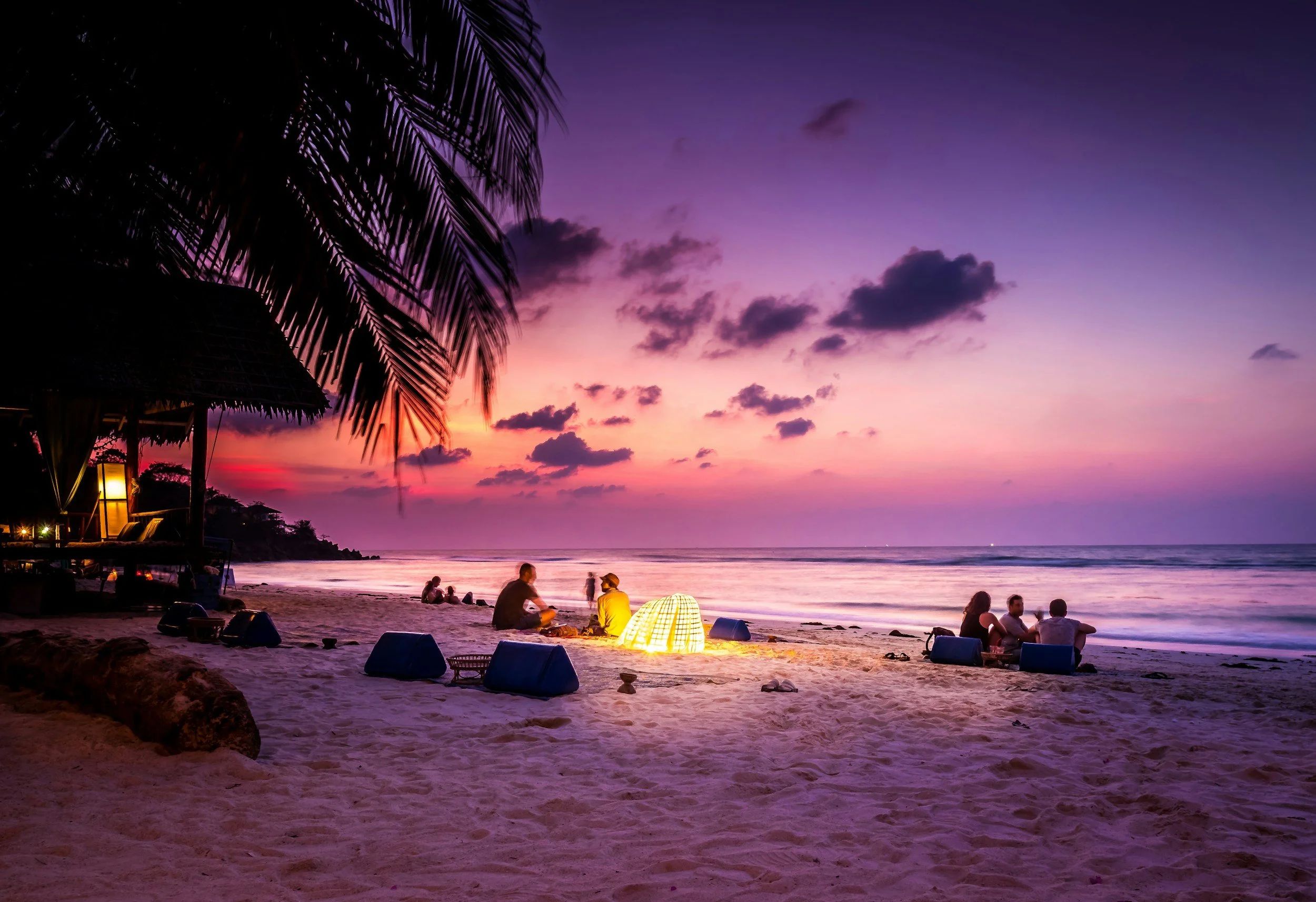 Several sets of people relax on an ocean beach at sunset. A palm tree on the left hangs over the beach.